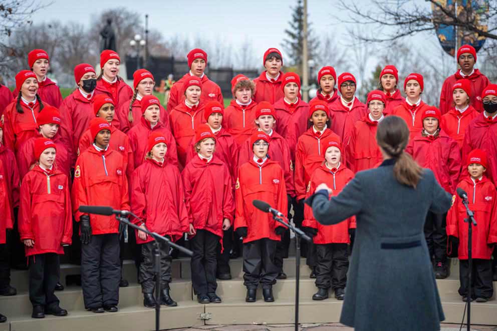 A children's choir sings the National Anthem, outdoors. They are all standing and wearing red clothing.