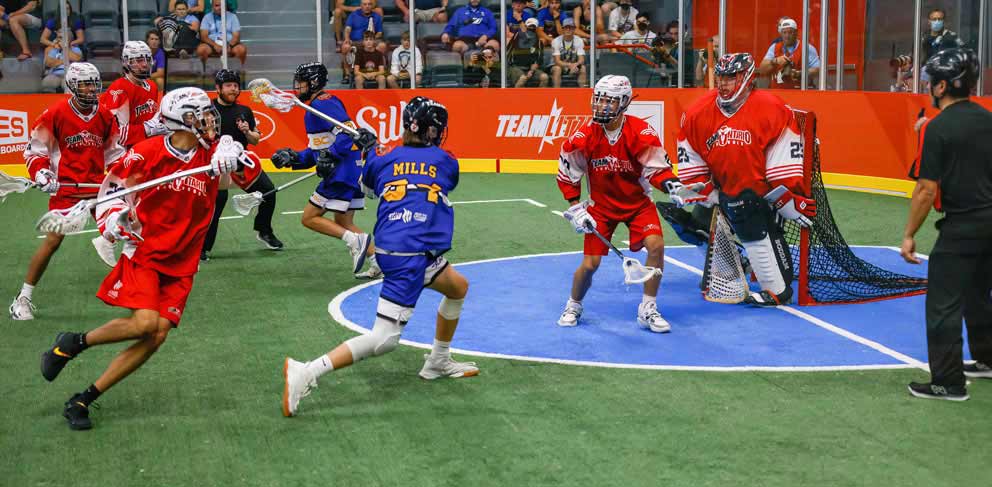 Players from two men's lacrosse teams compete during a match on an indoor surface.