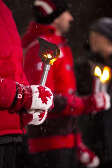 A person wearing a red coat and red and white maple leaf mittens holds a small torch during an outdoor wintertime celebration.