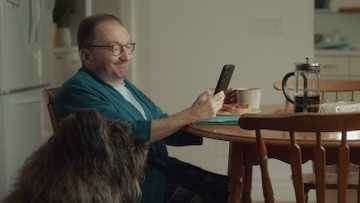 A person sits at their kitchen table to do their taxes, holding a mug in one hand and their cellphone in the other. There is a dog sitting next to them.