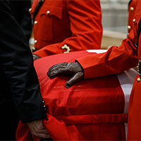 Close-up of the casket covered with the National Flag of Canada. We see the arm and upper body of an RCMP pallbearer in red serge whose black-gloved hand lie on the casket. 