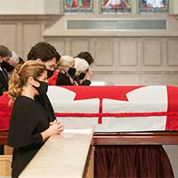 Le très honorable Justin Trudeau et Sophie Grégoire Trudeau sont agenouillés dans un banc d’église. On voit le cercueil, couvert du drapeau national du Canada, à l’arrière-plan.