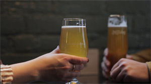 Photo of two pairs of hands holding full beer pints, with the word “Nonsuch” visible on one of the pint glasses