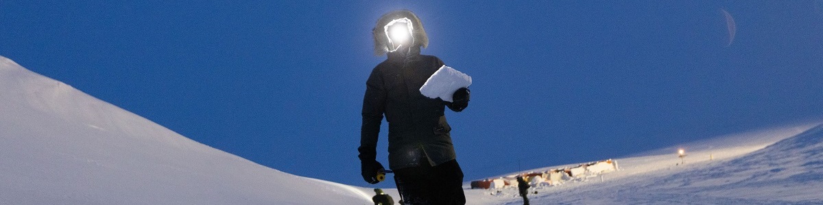 A person holding a rectangular piece of snow and wearing a headlamp stands in a snow covered field.