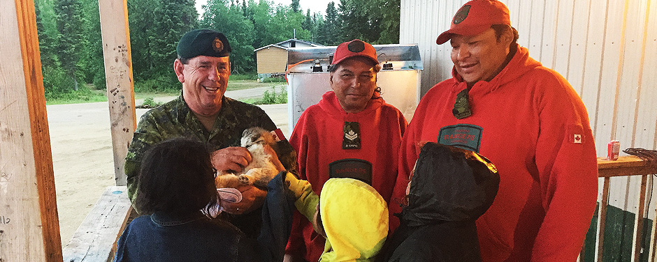 Slide - Warrant Officer Barry Borton, an army instructor, plays with a local puppy while talking to children in Wapekeka with Canadian Ranger Master Corporal Jacob McKay, and Sergeant Spencer Anderson