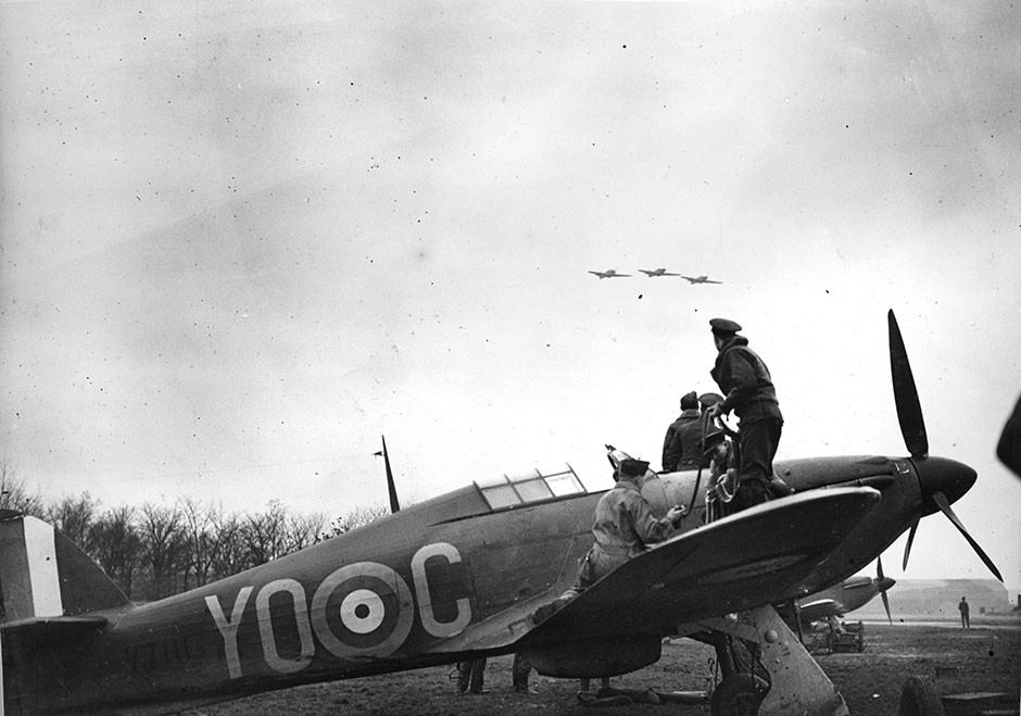 Photo de la bataille d¿Angleterre. Un Hurricane du 1er Escadron de l¿Aviation royale canadienne est réarmé pendant qu¿une section survole l¿aérodrome. L¿aéronef arbore l¿indicatif de l¿unité : « YO », Photo tirée de l¿album de photos du lieutenant d¿aviation R.W.G. Norris, de Saskatoon, en Saskatchewan, qui a servi dans le 1er Escadron pendant la bataille d¿Angleterre. PHOTO : Avec la permission du Musée national de la Force aérienne du Canada