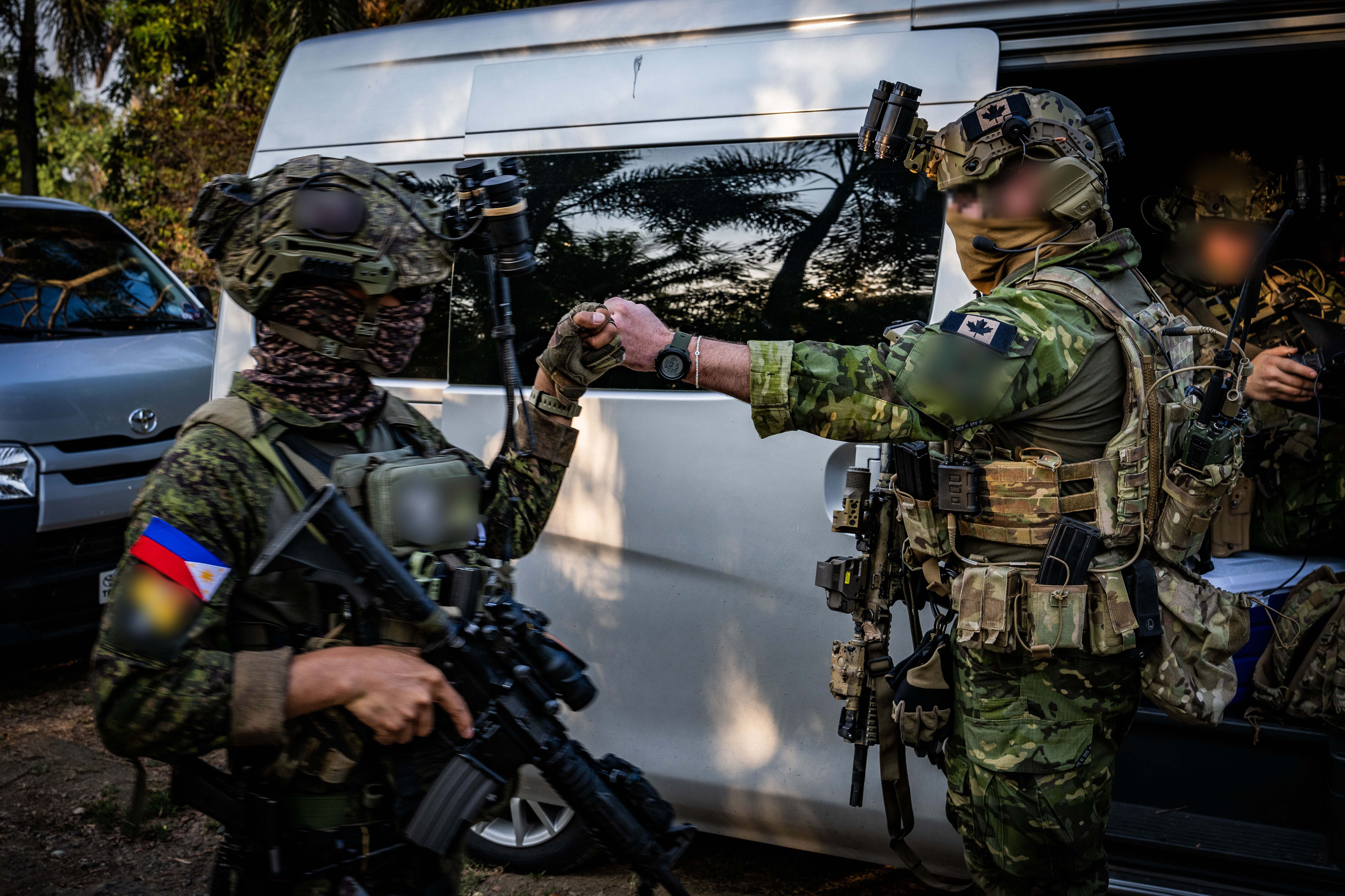 Two individuals wearing different uniforms and combat gear of various green shades, both with carbines and their faces covered with scarves, are fist bumping in front of a gray coloured minivan in a wooded area. One has a Philippines flag shoulder patch and the other has a Canadian flag shoulder patch.