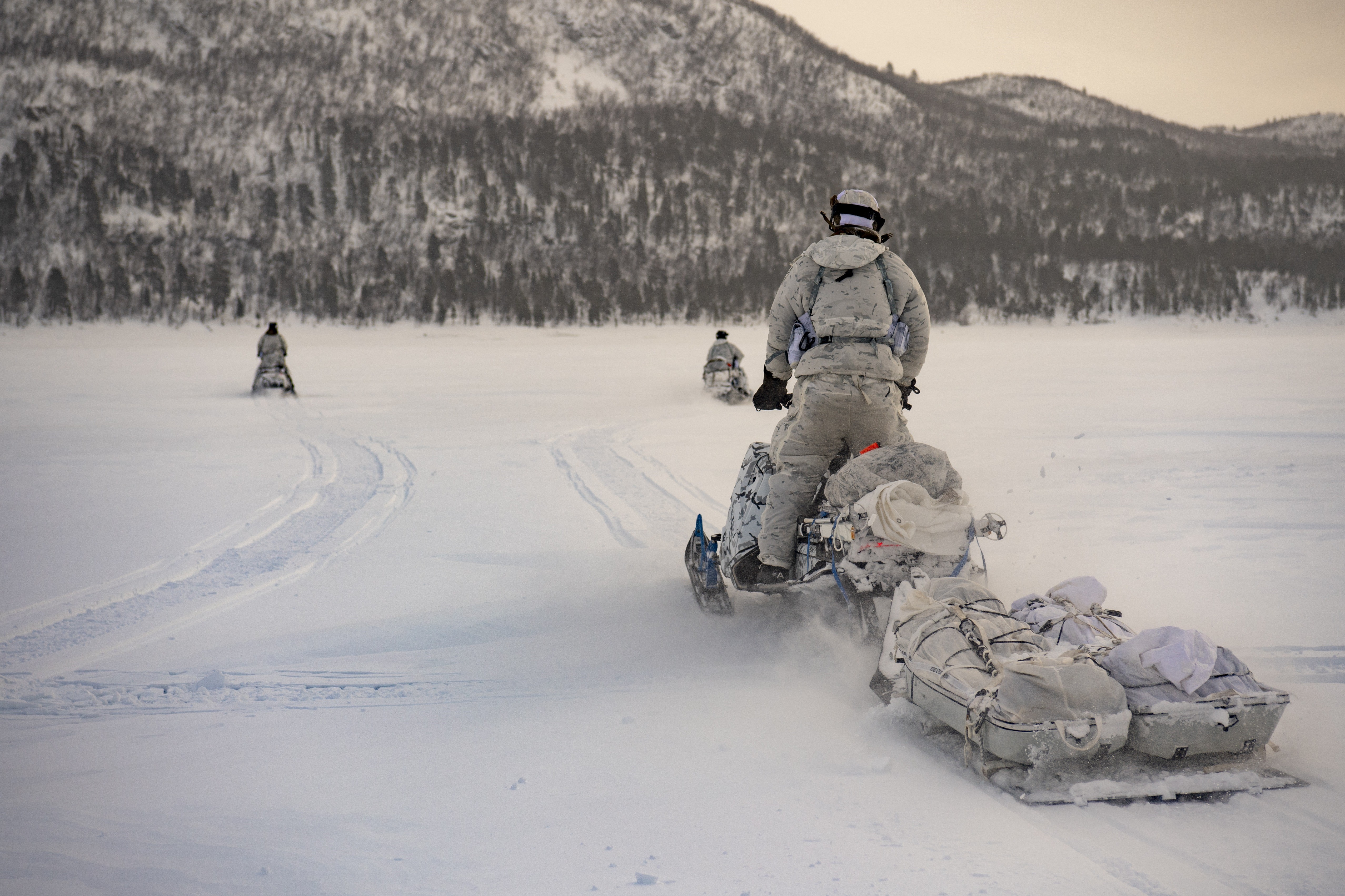Un militaire portant des vêtements de camouflage blancs utilise une motoneige en se tenant debout sur celle-ci pour voir plus loin. La motoneige tire une remorque contenant deux toboggans blancs sur une vaste surface plate enneigée vers des collines boisées. Deux autres militaires sur des motoneiges sont visibles plus loin devant le premier et se dirigent vers la même colline.
