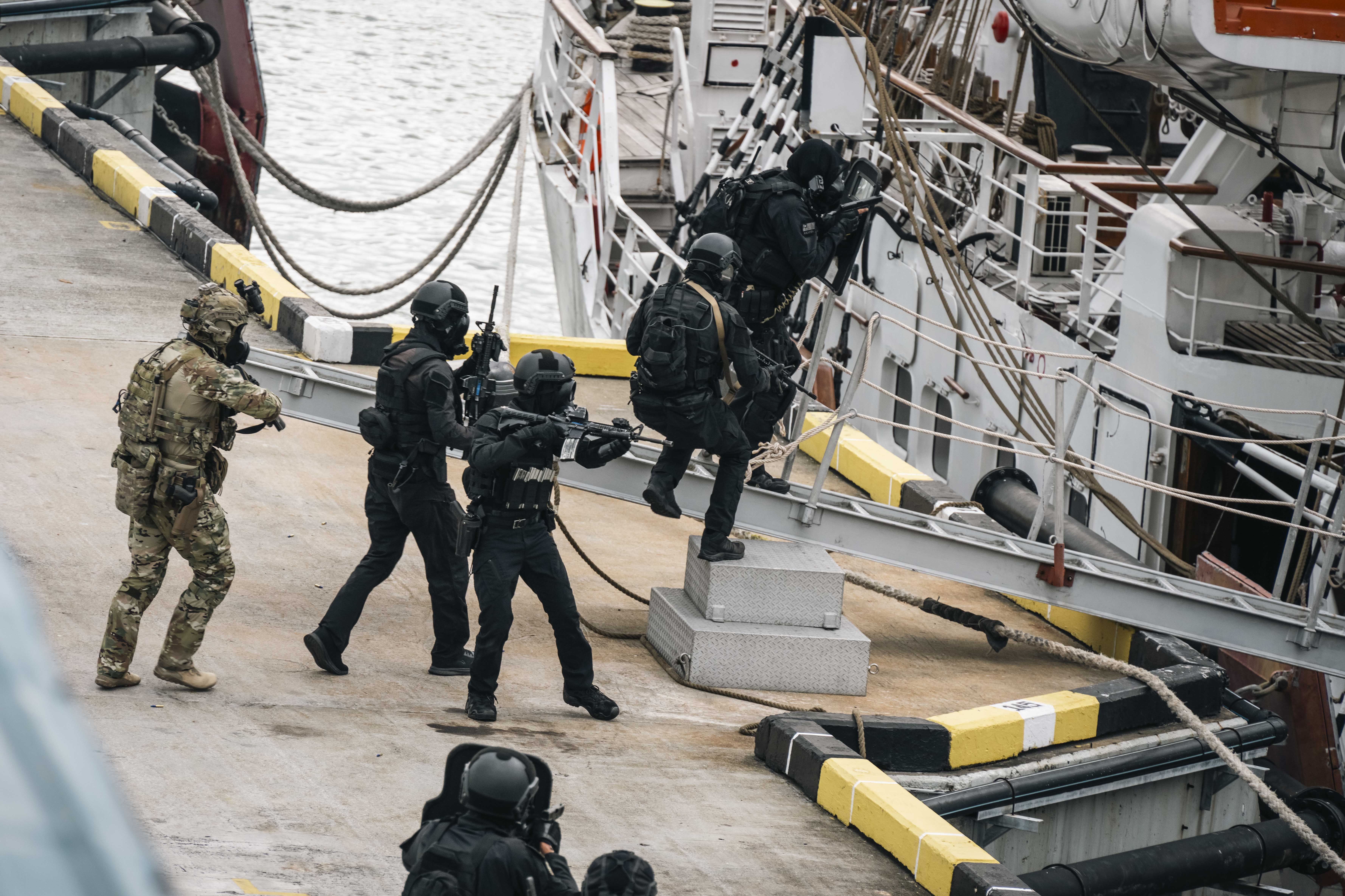 Quatre militaires malaisiens vêtus d’uniformes noirs et d’un équipement de combat, portant des masques à gaz et des carabines, montent sur la passerelle d’un navire à quai pour embarquer, suivis de près par un militaire canadien en uniforme Multicam, équipement de combat et masque à gaz. Le premier militaire malaisien tient un bouclier noir levé devant lui. Ils se trouvent sur un quai en béton au bord de la mer.