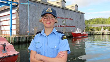 A woman in uniform stands smiling near a waterfront. Behind her is a building and a red boat on the water.