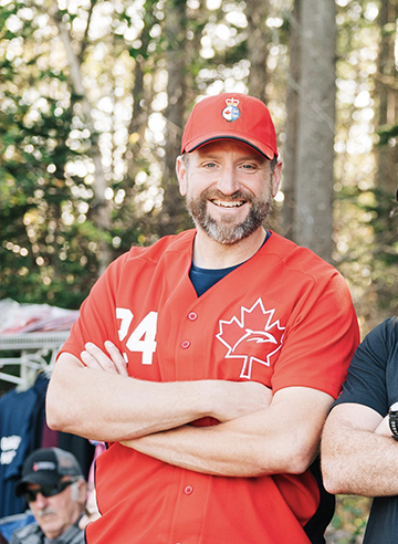Man stands outdoors, wearing a red baseball jersey and cap. His arms are crossed, and trees fill the background.
