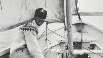 A man wearing a sweater and cap, sitting in a sailboat.