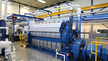 Large industrial engine in a factory setting, surrounded by pipes, gauges and metal flooring.