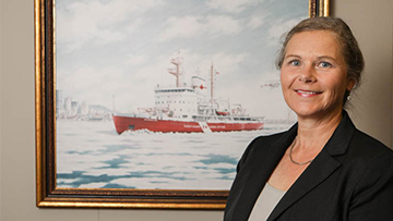 Woman in a suit stands smiling in front of a framed painting depicting a Canadian Coast Guard vessel at sea.