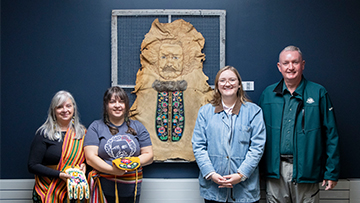 Four people stand in front of a wall with Indigenous artwork. Two individuals hold colourful beaded items.