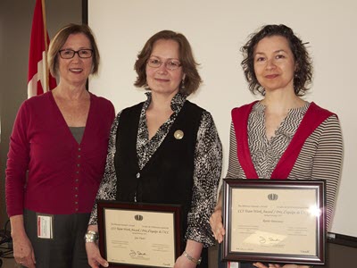 Three women smiling at the camera; two of them hold framed certificates.