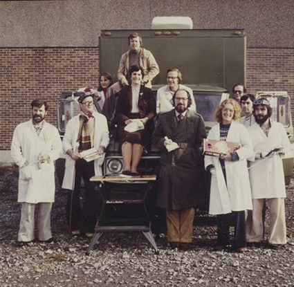 A photo of eleven conservators standing in front of and around the mobile lab. Most are wearing lab coats. Some are sitting on the hood and the roof.