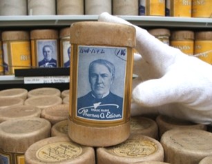 Une main gantée de blanc tient une petite cartouche sur laquelle on voit une photo de Thomas Edison