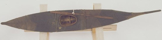 A model kayak made of dark brown sealskin with a paddle, displayed on a wooden stand.