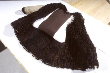 Manteau en peau de mouton brun foncé dont les longs poils sont à l'envers du vêtement. Un coussin rembourré est placé à l'intérieur.