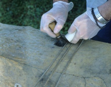 Detail showing the hands of a conservator protected with gloves. The conservator is using a brush and vacuum to remove green mould from the surface of a kayak.
