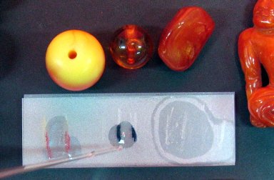 Trois perles : une ronde de couleur or, une ronde couleur rouge et une oblongue de couleur rouge. La lame en verre présente trois égratignures directement sous chaque perle. Une goutte de réactif est versée sur chaque égratignure.