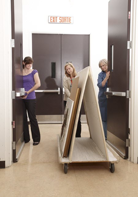 A person pushes an A-frame trolley through a set of double doors, which are held open by two assistants who act as spotters.