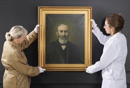Two conservators hold a framed, easel-sized painting in a vertical position against a wall.