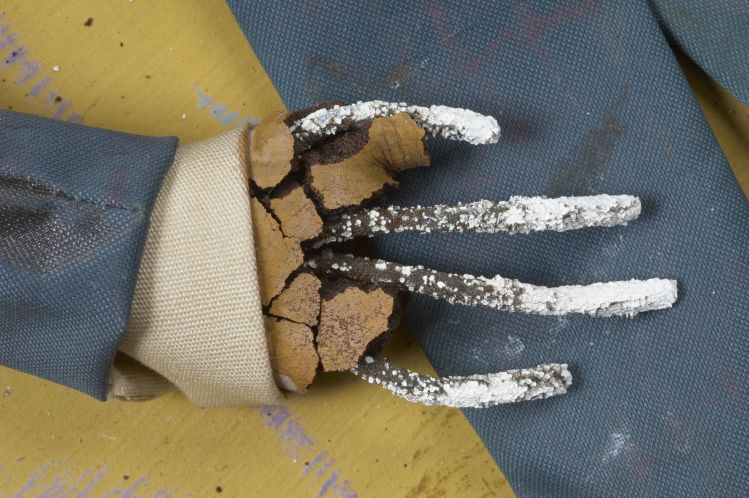 A puppet's hand extends from the cuff of a shirt or coat. The brown fabric that was used to make the hand has mostly disintegrated, revealing four fingers of gray metal. The metal is partly covered with white.