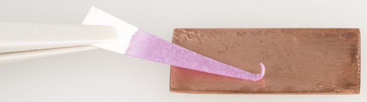 The tip of a pair of tweezers is holding a triangle of paper against a rectangle of copper metal. The end of the paper touching the copper is pink.