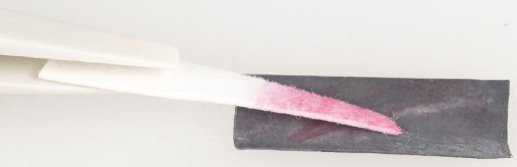 The tip of a pair of tweezers is holding a triangle of paper against a rectangle of lead metal. The end of the paper touching the lead is pink.