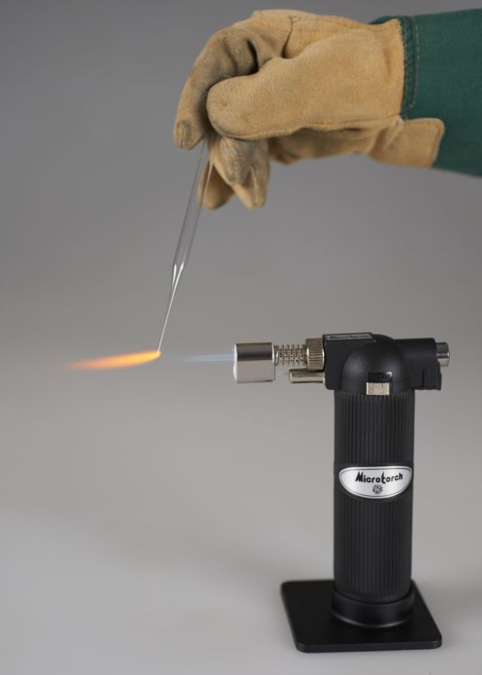 A gloved hand holds one end of a pipette so that the tip at the other end is in the flame of a butane torch.