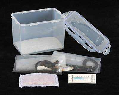 Container with the lid off, two polyethylene bags with zipper closures containing iron objects, silica gel and a humidity indicator card.