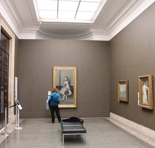 PMuseum visitor stands in a room with three paintings in fully enclosed frames.