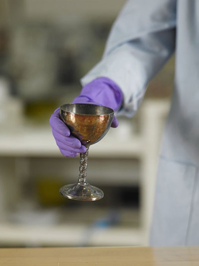 A hand wearing nitrile gloves is holding a tarnished silver-plated goblet.