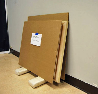 A painting placed on padded blocks on the floor next to a wall, in between two protective cardboard panels.
