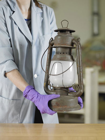 Gloved hands (nitrile gloves shown here) are used to carry an iron and glass lantern at its base.