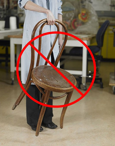 A chair being carried from the top of the back. A red circle with a diagonal line running through it covers most of the image and indicates that this is not appropriate.
