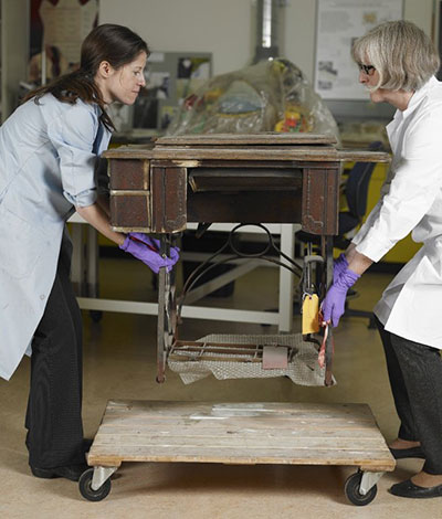 Une table de machine à coudre avec pédale est soulevée par sa base en fonte à l’aide de mains gantées.