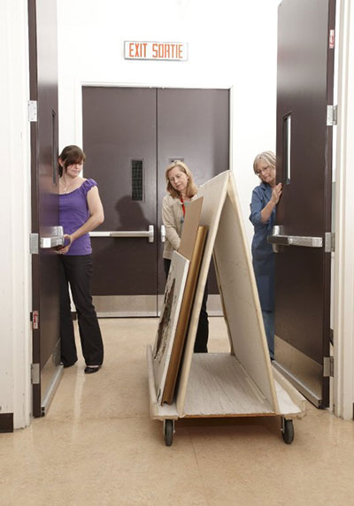 Framed and unframed paintings are moved on an A-frame cart. A cardboard sheet separates each piece.