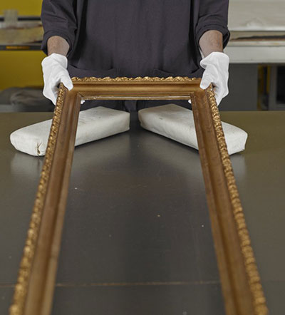 A gold-painted frame being lifted at one end by a handler wearing white gloves.