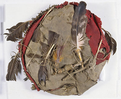 Buffalo hide warshield with red wool apron decorated with metal tinkle cones and feathers.