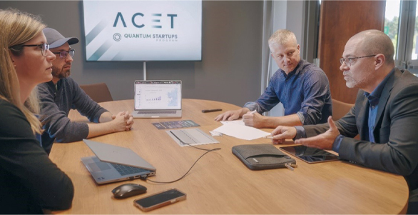 The photo shows a professional mentoring session for a start-up in the quantum field. Four people are sitting around a conference table.