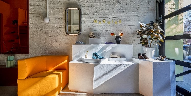 Présentoirs avec chaussures et objets décoratifs, un canapé à gauche et une plante en pot à droite, dans une boutique lumineuse au style minimaliste.