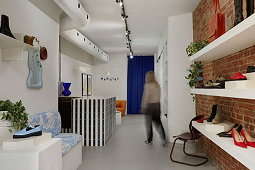 Intérieur d’une boutique avec étagères présentant des chaussures et sacs, un comptoir et une personne marchant vers le fond.