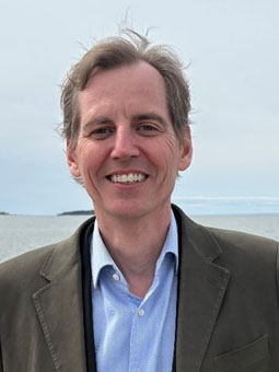 Portrait de Stéphane Arsenault, directeur régional du bureau d’affaires Bas-Saint-Laurent, debout devant un plan d’eau, portant une veste et une chemise, avec un paysage maritime et un ciel dégagé en arrière plan.