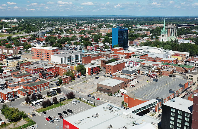 Aerial view of downtown Trois-Rivières featuring commercial and residential buildings, grid-patterned streets and parking areas, under a partly cloudy sky.