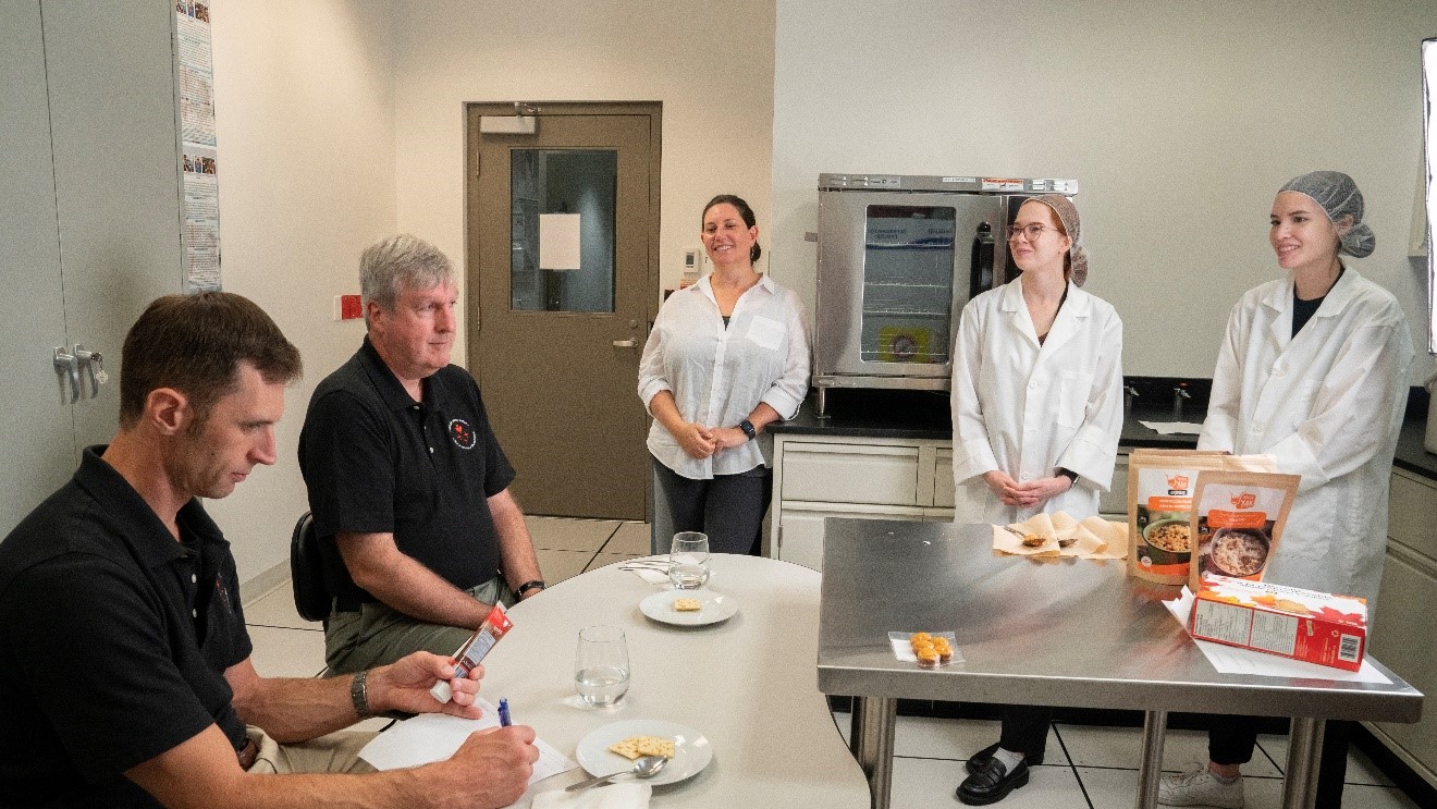 L’astronaute Joshua Kutryk teste des repas lyophilisés Happy Yak dans un laboratoire alimentaire en présence de quatre personnes.