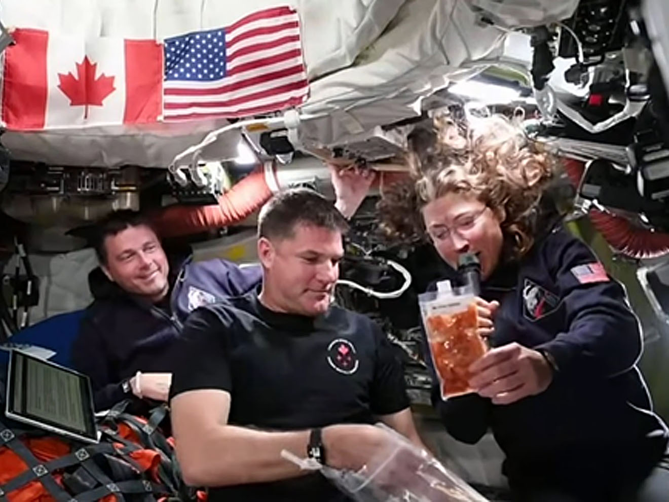 Les astronautes Reid Wiseman, Jeremy Hansen et Cristina Koch manipulent des sachets de nourriture à bord de la capsule Orion, sous des drapeaux canadien et américain, entourés d’équipements techniques.