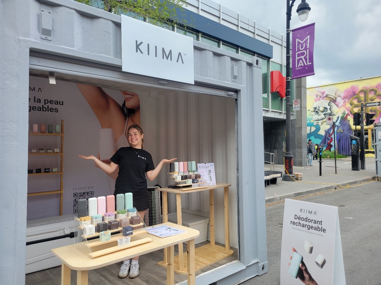 Kiima booth set up in an urban container, showcasing refillable deodorants and personal care products displayed on tables and shelves, with a person behind the counter and a promotional sign at the front.